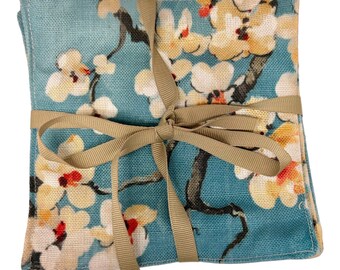 May include: Set of four blue and white floral fabric coasters tied together with a brown ribbon. The coasters feature a floral pattern with white flowers and brown branches on a blue background.