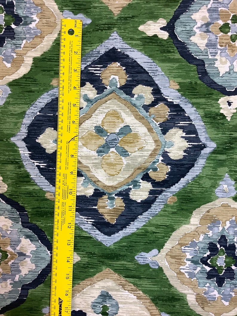 May include: Green, blue, and beige fabric with a repeating pattern of stylized flowers and geometric shapes. The fabric is measured with a yellow ruler.