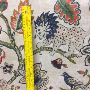 May include: A beige fabric featuring a multi-colored pattern of lions, birds, and floral motifs. A yellow ruler is placed vertically on the fabric, showing a measurement scale of 12 inches, indicating the scale of the printed design elements.