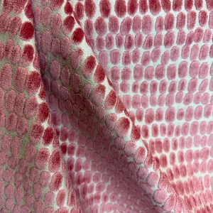 May include: Close-up of a pink and red patterned fabric. The fabric features a repeating pattern of raised, oval-shaped designs in varying shades of pink and red against a white background. The fabric is folded, showing its texture and depth.