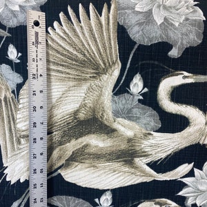 May include: Black fabric with a white and gray crane design. The crane is flying with its wings spread. The background is a dark black with a subtle texture. There are also white and gray lotus flowers in the background.