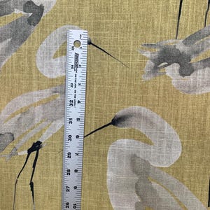 Pode incluir: Close-up de um tecido com um desenho de guindaste em aquarela em cinza e preto sobre um fundo amarelo dourado. Uma r&eacute;gua mostra as medidas em polegadas, cerca de 61-89 cm.