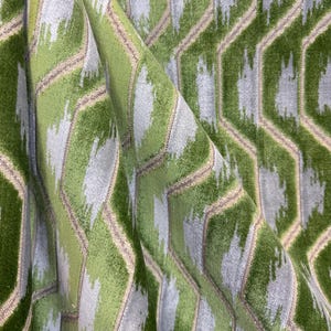 May include: Close-up of a textured fabric with a geometric pattern. The design features a repeating pattern of green and gray shapes outlined with a tan color. The fabric is folded, showing the texture and depth of the material.