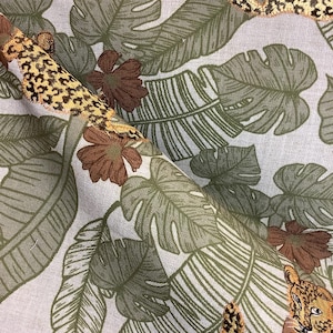 May include: Close-up of a fabric with a tropical jungle theme. The fabric features large green leaves, brown flowers, and embroidered leopard print accents. The design includes a leopard's face and tail, adding a playful touch to the botanical pattern.