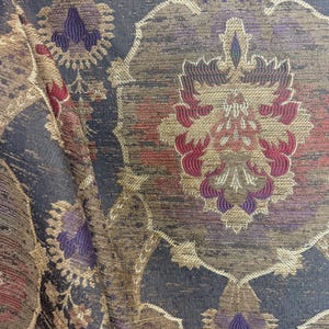 May include: Close-up of a patterned fabric with a dark blue background and ornate floral designs in gold, red, and purple. The fabric has a textured appearance, with a slight sheen. The design is reminiscent of vintage textiles.