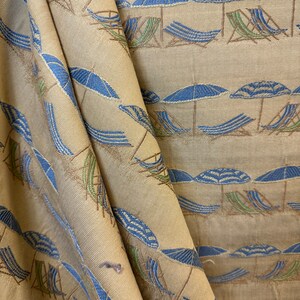 May include: Beige fabric with a repeating pattern of blue and white striped beach umbrellas and brown deck chairs. The fabric is folded in half, creating a diagonal line.