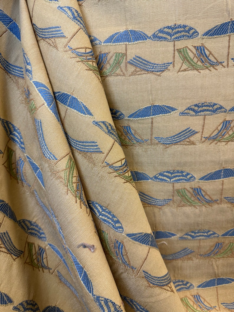 May include: Beige fabric with a repeating pattern of blue and white striped beach umbrellas and brown deck chairs. The fabric is folded in half, creating a diagonal line.