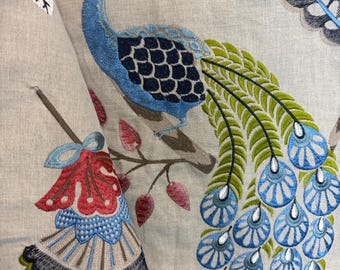 May include: Beige fabric with an embroidered peacock design. The peacock is blue, green, and black, perched on a brown branch with red and pink leaves. The fabric has a detailed, textured appearance, suitable for home decor.