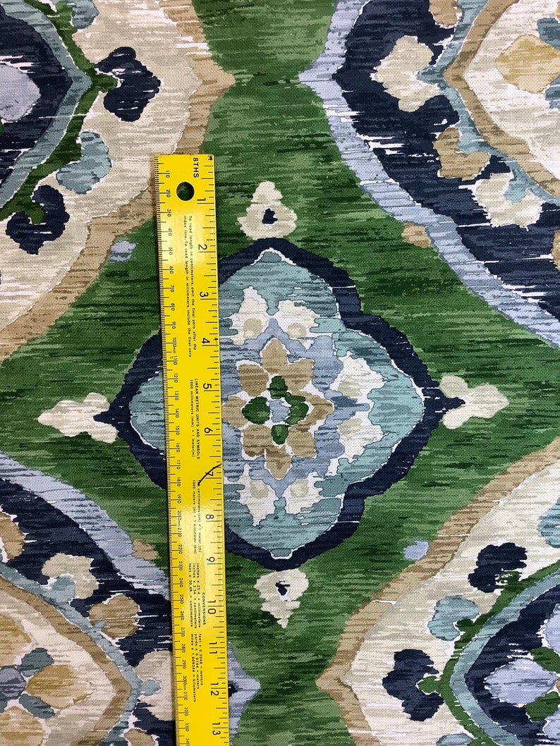 May include: A close-up of a fabric with a repeating pattern of blue, green, and beige. The fabric has a textured, brushed look. A yellow ruler is placed on the fabric to show the scale.