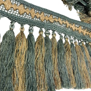 May include: Close-up of decorative trim with teal and gold tassels. The trim features a teal base with a gold, wavy design. The tassels are a mix of teal and gold, adding a touch of elegance. Ideal for home decor and craft projects.