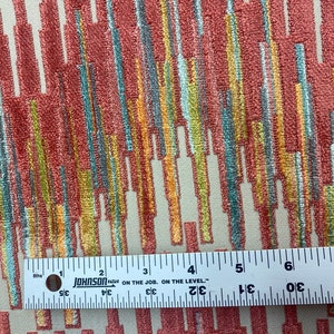 Op de afbeelding: Een close-up van een stof met een kleurrijk abstract patroon. De stof heeft een textuur, bijna een pluche oppervlak. Het patroon bestaat uit verticale lijnen in verschillende kleuren, waaronder rood, oranje, geel, groen en blauw. Een liniaal ligt bovenop de stof, wat aangeeft dat de stof ongeveer 15 cm breed is.