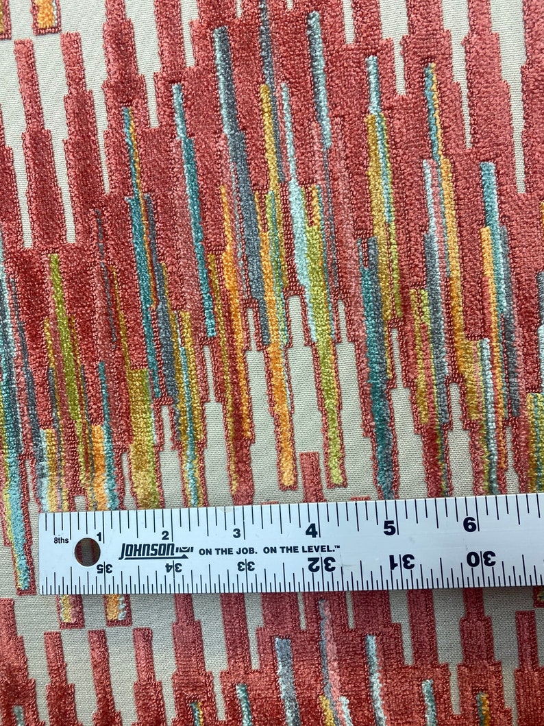 Op de afbeelding: Een close-up van een stof met een kleurrijk abstract patroon. De stof heeft een textuur, bijna een pluche oppervlak. Het patroon bestaat uit verticale lijnen in verschillende kleuren, waaronder rood, oranje, geel, groen en blauw. Een liniaal ligt bovenop de stof, wat aangeeft dat de stof ongeveer 15 cm breed is.