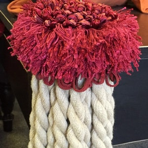 May include: A close-up of a decorative tassel with a red and gold rope and a white braided tassel. The tassel is hanging from a dark wooden post.