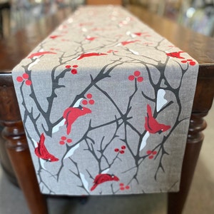 May include: A gray linen table runner with a wintery design featuring red cardinals perched on branches with red berries.