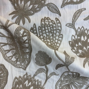 May include: Close-up of white fabric with embroidered floral design in shades of taupe. The pattern features large, stylized flowers, leaves, and stems. The fabric has a subtle textured appearance, with visible vertical lines.