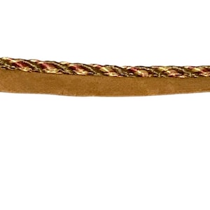 May include: Close-up of a decorative trim with a braided rope design in gold, red, and cream colors. The trim is attached to a tan suede-like material, creating a textured and elegant look. Ideal for crafting and sewing projects.