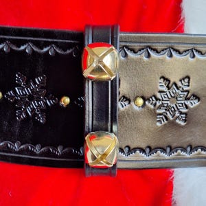 May include: A black leather belt with gold-tone metal bells and snowflake designs. The belt is decorated with a scalloped edge and has a buckle.