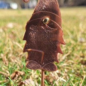 May include: A copper gnome garden stake with a gold eye detail. The gnome is standing in a grassy area.