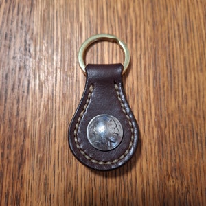 May include: A brown leather keychain with a stitched edge and a silver buffalo nickel coin inset. The coin has the year 1957 on it.