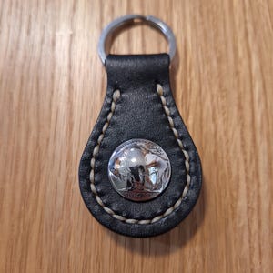 May include: Black leather keychain with a silver nickel coin featuring a buffalo and the words "FIVE CENTS." The keychain has a silver key ring and is stitched with cream-colored thread. A classic and durable accessory.