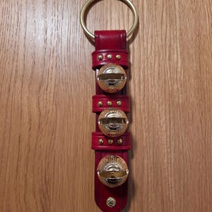 May include: A red leather door hanger with three gold jingle bells. The hanger has a gold ring at the top for hanging.