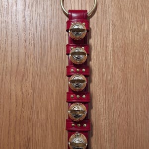 May include: A red leather jingle bell door hanger with six gold bells. The bells are attached to the leather strap with a gold ring at the top.