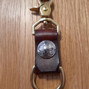 May include: A leather key fob with a brass clasp and ring. The key fob features a brown leather strap with a silver coin displaying a buffalo. The metal hardware is gold-toned.