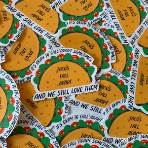 K&ouml;nnte beinhalten: Eine Sammlung bunter Aufkleber mit einem Cartoon-Taco mit dem Text "Tacos Fall Apart" und "And We Still Love Them". Die Aufkleber sind auf wei&szlig;em Papier gedruckt und haben eine gl&auml;nzende Oberfl&auml;che.