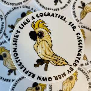 May include: A circular white sticker with a yellow cockatiel design. The bird is depicted on a branch, with a black beak and brown eyes. The text around the edge says: "She's like a cockatiel, sir. Fascinated by her own reflection."