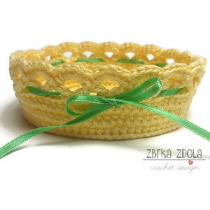 May include: A yellow crocheted basket with a green ribbon tied around it. The basket is on a white background. The text "ZBRKA ZDOLA crochet design" is visible below the basket.