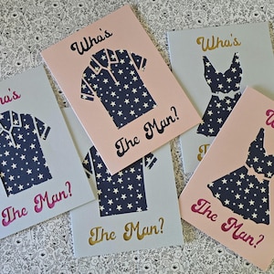 May include: Greeting cards with cut-out designs of a shirt and a dress, both with a navy blue star pattern. The cards are in shades of pink and gray, with the text "Who's The Man?" in various colors.