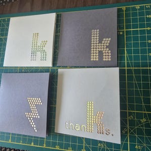 May include: Four square greeting cards with gold dot designs. One card features the letter "k", another a lightning bolt, and a third the word "thanks". The cards are in shades of gray and off-white.