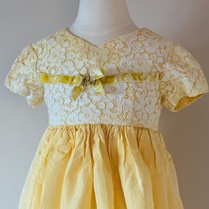 May include: A yellow and white lace dress with a yellow velvet bow. The dress has short sleeves and a full skirt.