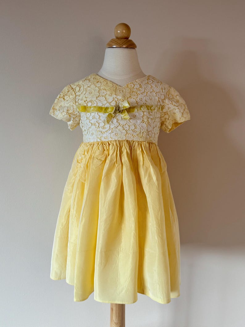 May include: A yellow and white lace dress with a yellow ribbon bow at the neckline. The dress has short sleeves and a full skirt.