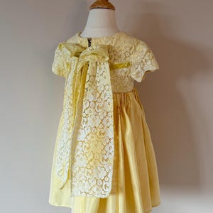 May include: A yellow lace dress with a large bow in the back. The dress has short sleeves and a full skirt.
