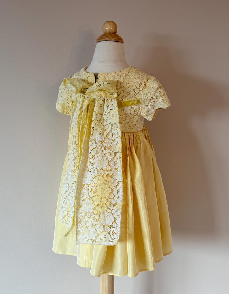 May include: A yellow lace dress with a large bow in the back. The dress has short sleeves and a full skirt.