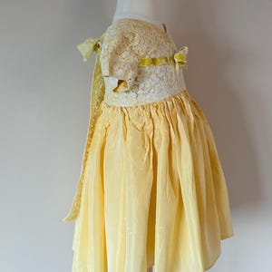 May include: A yellow lace and silk dress with a yellow velvet ribbon bow. The dress has a gathered skirt and short sleeves.