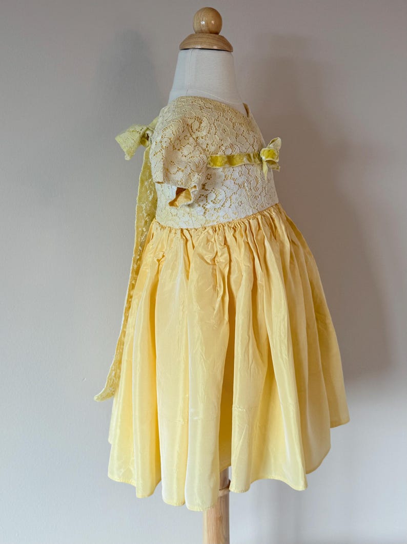 May include: A yellow lace and silk dress with a yellow velvet ribbon bow. The dress has a gathered skirt and short sleeves.