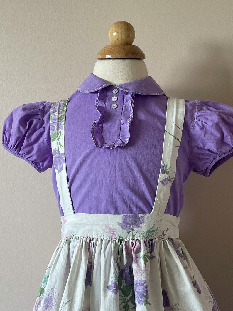 May include: A purple collared blouse with puffed sleeves and a white ruffle collar, worn over a white skirt with a floral print and suspenders.