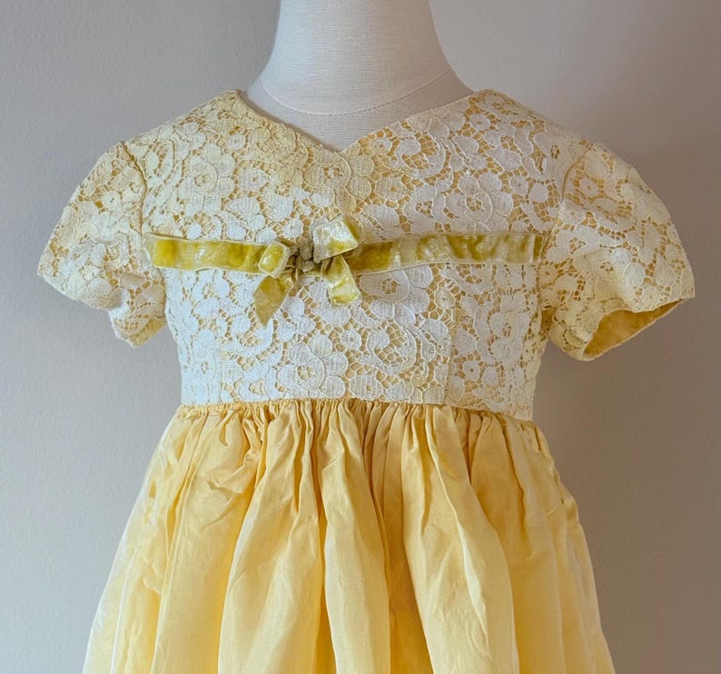May include: A yellow and white lace dress with a yellow velvet bow. The dress has short sleeves and a full skirt.