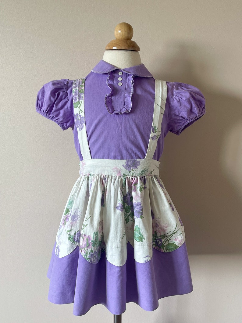 May include: A purple and white dress with a floral print. The dress has a white bodice with purple floral accents and a purple skirt with a scalloped hem. The dress is on a mannequin.