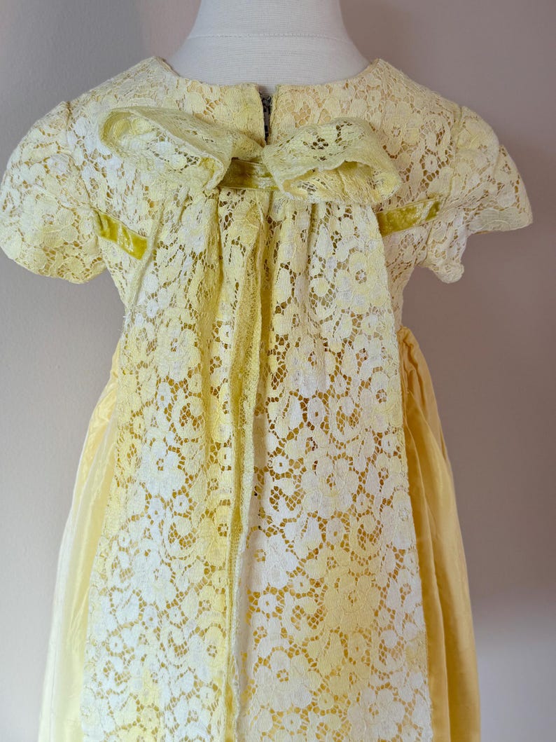 May include: A yellow lace dress with a large bow in the back. The dress has a fitted bodice and a full skirt.