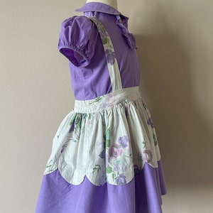 May include: A purple and white floral print dress with a ruffled collar and straps. The dress has a scalloped hem and is made of a soft, lightweight fabric.