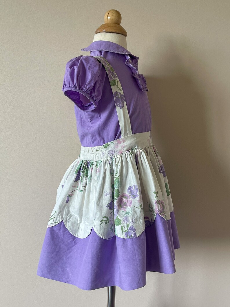 May include: A purple and white floral print dress with a ruffled collar and straps. The dress has a scalloped hem and is made of a soft, lightweight fabric.