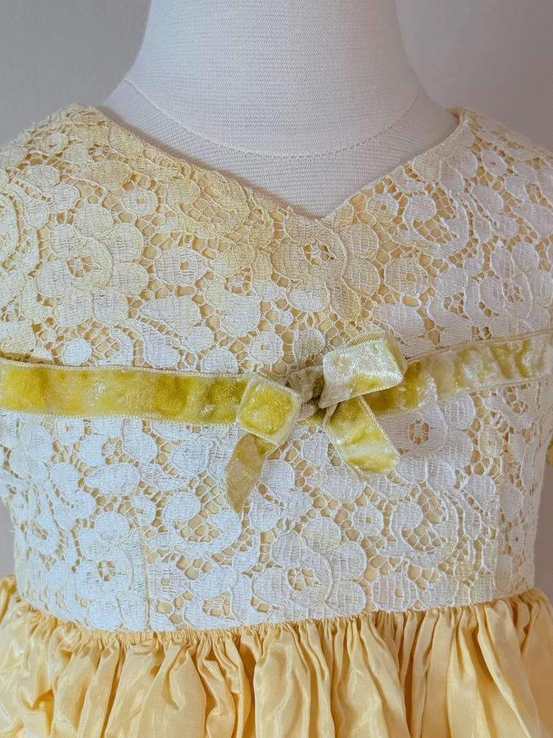 May include: A yellow lace dress with a velvet bow at the neckline. The dress has a ruffled skirt and is made of a soft, delicate fabric.