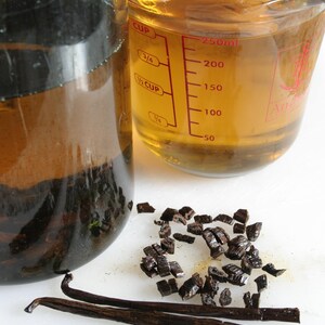 May include: A glass jar filled with a dark liquid and vanilla beans, a measuring cup filled with a light yellow liquid, and a pile of vanilla bean pieces on a white surface.