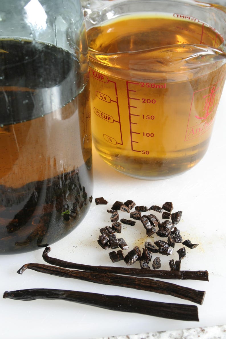 May include: A glass jar filled with a dark liquid and vanilla beans, a measuring cup filled with a light yellow liquid, and a pile of vanilla bean pieces on a white surface.