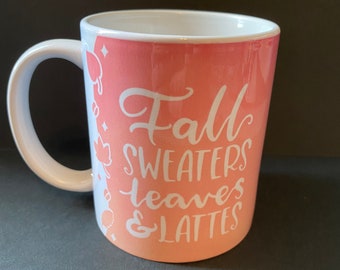 May include: A white ceramic mug with a pink ombre design. The mug has the text "Fall sweaters leaves & lattes" in white lettering.