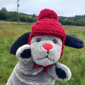 May include: A plush grey dog hand puppet featuring long black ears, a bright red nose, and a red knitted hat with a pom-pom on top. The puppet has orange eyes and a small red fabric bow tied around its neck, with black accents on its paws.