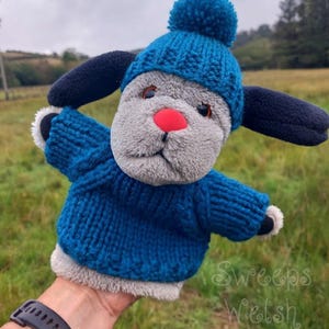 Könnte beinhalten: Eine Plüsch-Hundehandpuppe mit grauem Fell, einer leuchtend roten Nase und dunkelbraunen Augen. Die Puppe trägt einen blauen Strickpullover und eine passende blaue Strickmütze mit einem Bommel oben. Lange dunkelblaue Ohren ragen seitlich hervor.